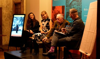 Czterech siedzących panelistów na scenie: z lewej dwie kobiety (jedna z długimi brązowymi włosami w ciemnym ubraniu, druga w czarno-białej sukience), obok starszy mężczyzna z siwymi włosami trzymający mikrofon, po prawej mężczyzna w garniturze z mikrofonem i notesem; w tle pionowy ekran z czarno-białym zdjęciem oraz niebiesko-czerwony plakat z napisem Archiwa Państwowe.