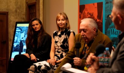 Trzy osoby siedzą przy panelu: po lewej długowłosa kobieta w czarnej bluzce, obok kobieta w sukience w czarno‑białe wzory ze splecionymi dłońmi, na pierwszym planie po prawej starszy mężczyzna trzyma mikrofon; w tle ekran z czarno‑białym zdjęciem, kolorowy pomarańczowo‑niebieski plakat i wzorzysta tapeta sali.