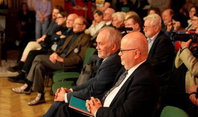 Widok z boku na rząd siedzących osób w sali: z przodu kilku starszych mężczyzn w garniturach i marynarkach na zielonych krzesłach z złożonymi rękami i notesami, w tle rozmyta publiczność i osoba trzymająca duży aparat fotograficzny.
