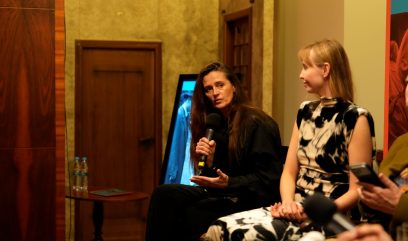 Dwie kobiety siedzące na panelu; po lewej kobieta z długimi ciemnymi włosami trzyma mikrofon, po prawej blondynka w czarno‑białej sukience patrzy w jej stronę, w tle drewniane drzwi, monitor z niebieskim ekranem i stolik z butelkami wody, z przodu rozmyte ręce innej osoby trzymającej telefon i mikrofon.