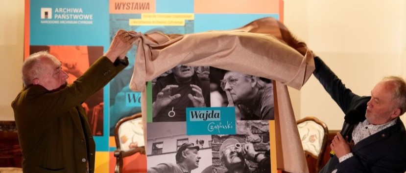 Dwaj starsi mężczyźni odsłaniają beżowym materiałem stojący plakat z kilkoma czarno-białymi zdjęciami i niebiesko-zielonym napisem Wajda, jeden z nich trzyma mikrofon; w tle kolorowe panele z napisami Archiwa Państwowe i Wystawa oraz krzesła.